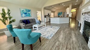 A spacious living room with a grey sofa, teal armchairs, and a coffee table. A kitchen and dining area are visible in the background.