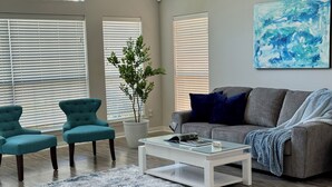 A living room with a grey sofa, two teal armchairs, a white coffee table, and a blue abstract painting on the wall.