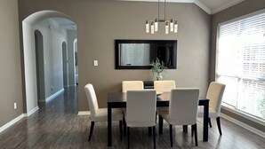 A dining area with a dark wood floor, a rectangular table with six chairs, a modern chandelier, and a large window with blinds.