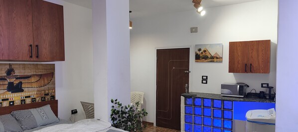 Une chambre à coucher comprenant un lit, des armoires en bois, un tableau représentant une scène égyptienne, une porte et un coin cuisine équipé d’un four à micro-ondes et d’un grille-pain.