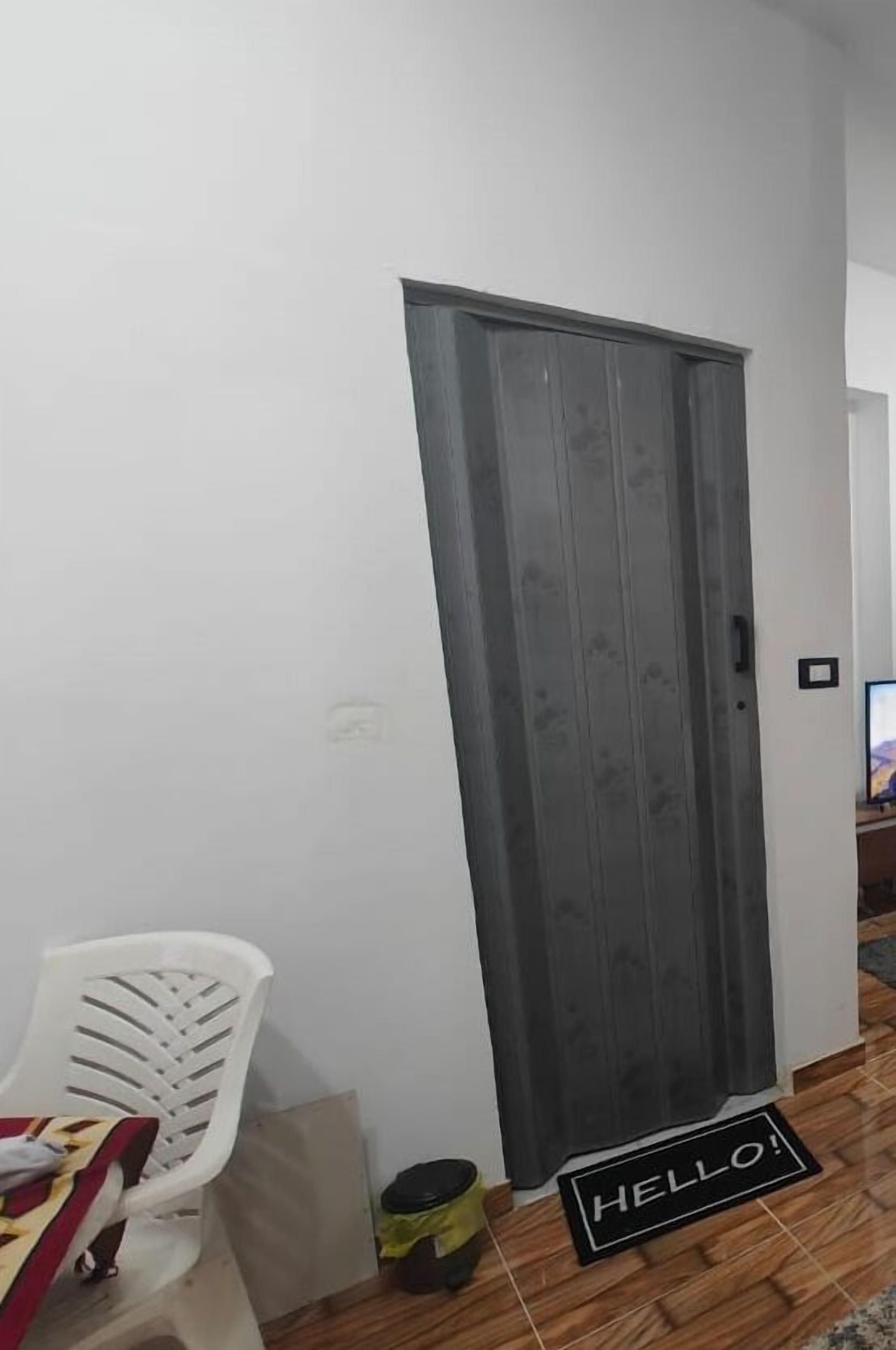A room with a grey door, a white plastic chair, and a 'HELLO!' mat on the floor.