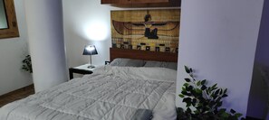 En snyggt bäddad säng med ett vitt täcke, en sänggavel i trä med ett konstverk med egyptiskt tema, ett litet nattduksbord med en lampa och en krukväxt till höger.