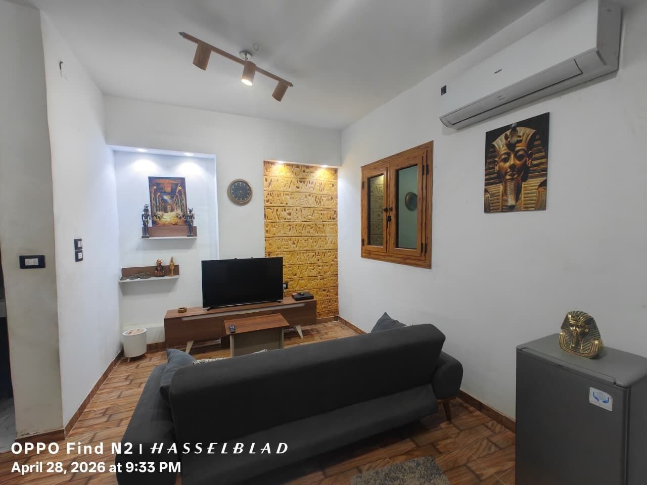 Sala de estar com sofá, TV, painéis de madeira, arte com temática egípcia e um pequeno armário.