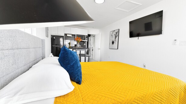 A hotel room with a bed, a grey headboard, a yellow quilted bedspread, a blue pillow, a flat-screen TV mounted on the wall, and a kitchen area visible through an open door.