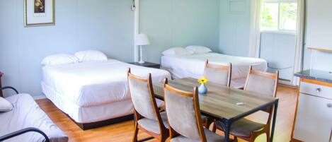 A room with two single beds, a wooden dining table with chairs, a small kitchen area, and a window with curtains.