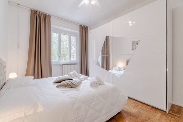 A modern bedroom with a large bed, a window with curtains, a mirror, and a wooden floor.