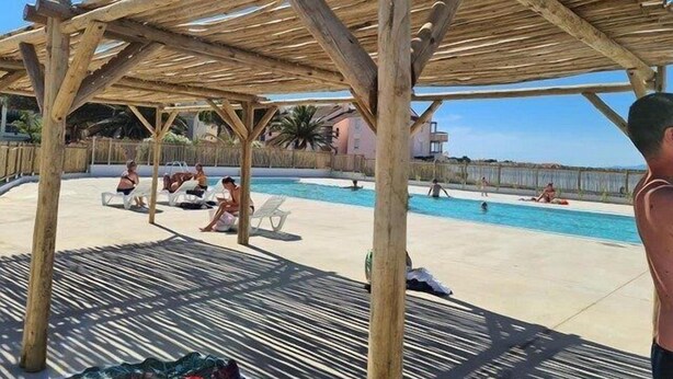 Une pergola en bois surplombe un espace piscine où des personnes se détendent et prennent des bains de soleil.