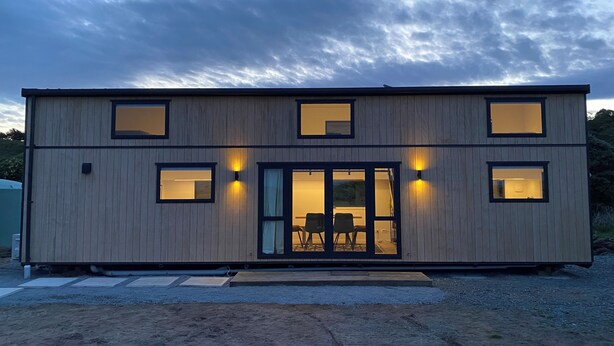 A modern two-story building with a wooden exterior, large windows, and a glass door leading to an interior with a seating area.