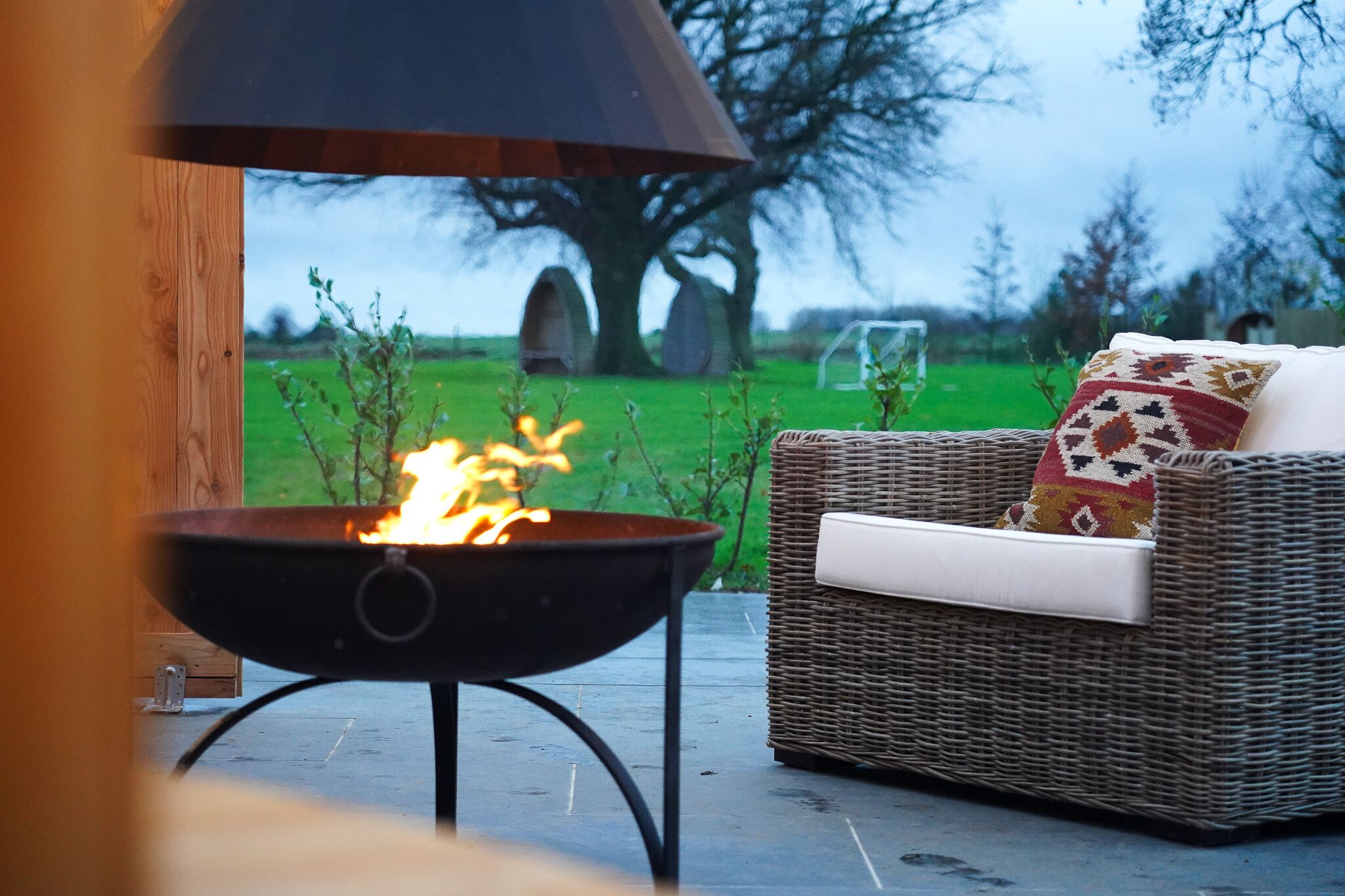 Un foyer avec des flammes, un canapé en osier avec un coussin, et une vue sur un champ verdoyant avec des arbres en arrière-plan.