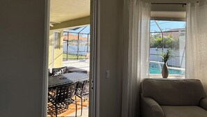 Ein Zimmer mit einer Schiebetür und Glasfront, die auf eine Terrasse mit Tisch und Stühlen führt. Im Zimmer befindet sich eine Couch und ein Fenster mit Vorhängen.