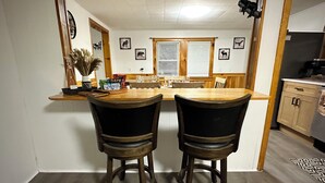 A dining area with two wooden chairs, a wooden table, and a kitchen in the background. - 4BR Summer Home For 10ppl with Game Room -White Mtns! (Woodstock)