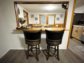 Uma área de refeições com duas cadeiras de madeira, uma mesa de madeira e uma cozinha ao fundo.