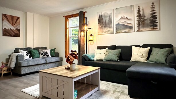 A living room with a grey sofa, a black sofa, a wooden coffee table, and a white side table. There are three paintings on the wall and a window with curtains. - 4BR Summer Home For 10ppl with Game Room -White Mtns! (Woodstock)
