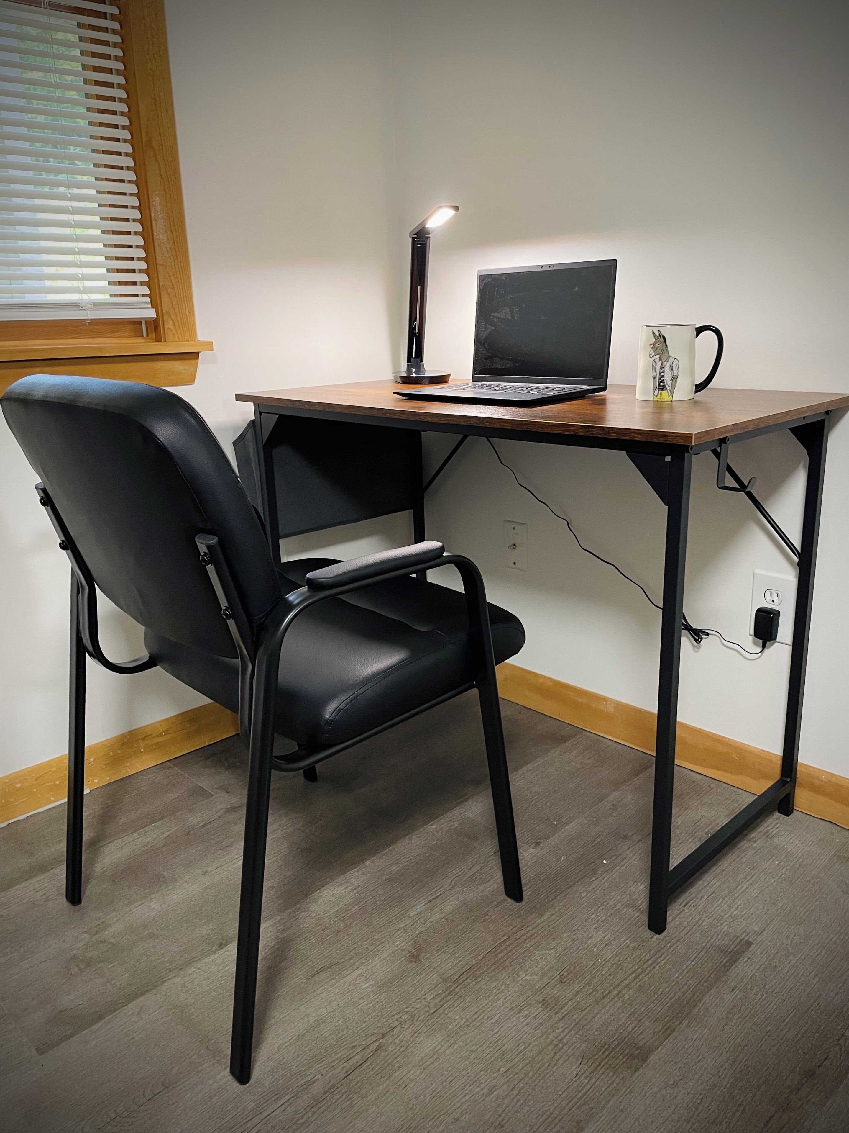 Ein Arbeitsplatz mit einem Holztisch, einem schwarzen Stuhl, einem Laptop, einer Schreibtischlampe und einer Tasse.