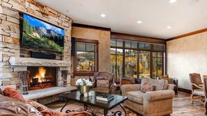 A living room with a stone fireplace, a flat-screen TV mounted on the wall, and a large window offering a view of trees and mountains. - Abode at Silver Summit, ski-in/ski-out, hot tub, pool table, Silver Lake (Park City)