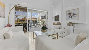 Een moderne woonkamer met witte banken, een glazen salontafel en uitzicht op een terras met een zwembad en palmbomen.