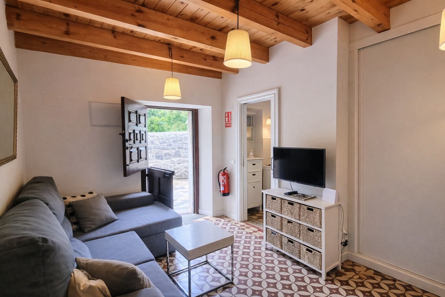 Een moderne woonkamer met een grijze bank, een klein wit tafeltje, een televisie op een wit meubelstuk en een vloer met een patroon.