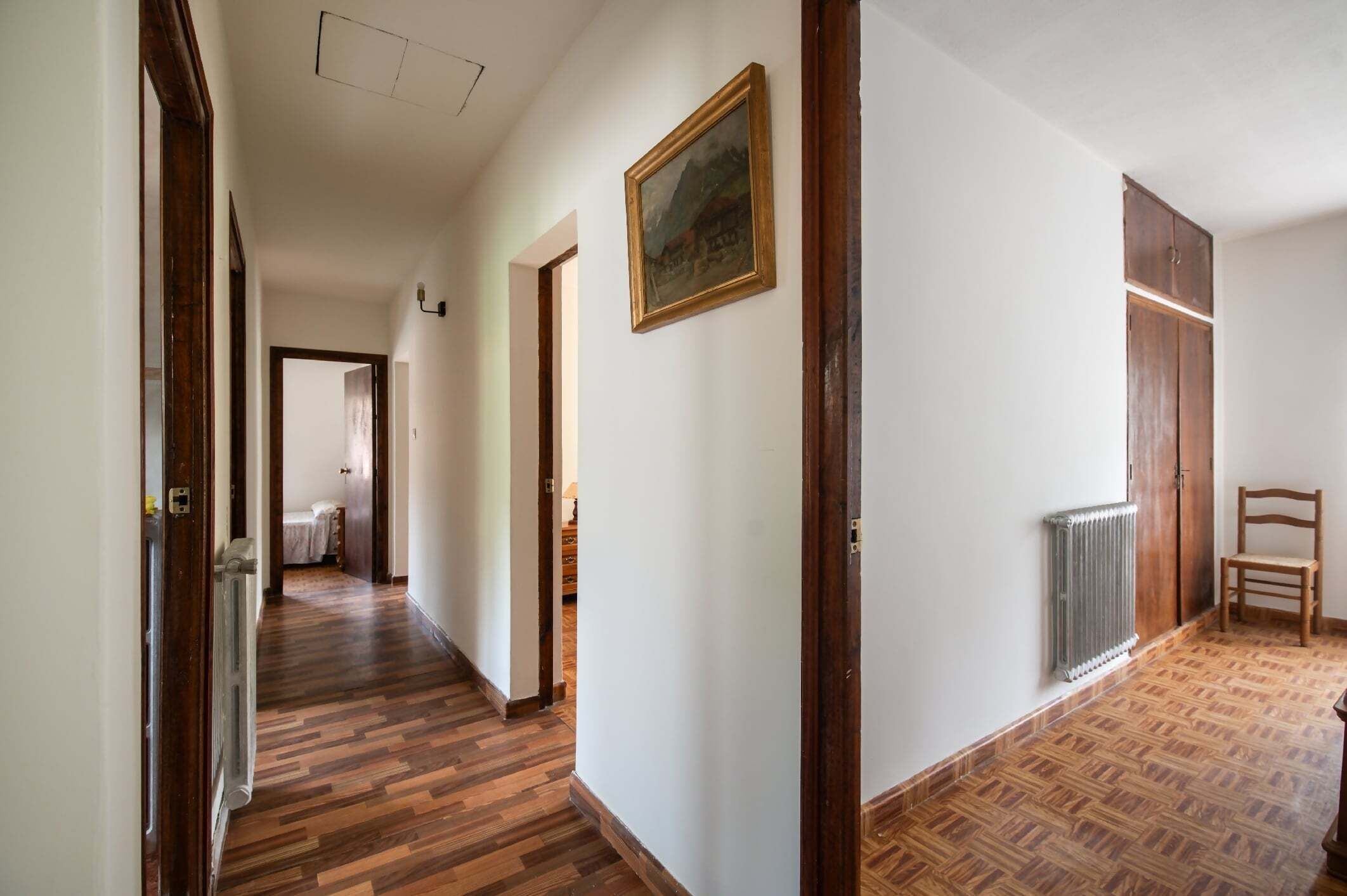 Un pasillo con suelo de madera, un radiador, una silla y un cuadro en la pared.