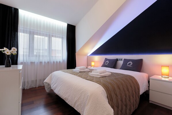 A modern hotel room with a large bed, a bedside table, a nightstand, and a window with sheer curtains.