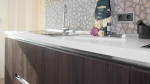 Una cocina moderna con encimera blanca, armarios de madera oscura, salpicadero de piedra y cafetera.