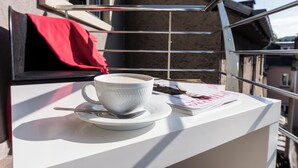 Un balcón con una mesa blanca, una taza blanca y su platillo, una cuchara, una revista y una toalla roja.