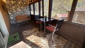 Een gezellige kamer met een stenen muur, een houten bank, een tafel met stoelen en grote ramen met uitzicht op bomen en een gebouw.