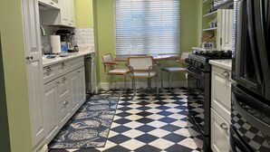 A compact kitchen with white cabinets, a black and white checkered floor, a dining area with two chairs, and a window with blinds. - Charming 3-bedroom apartment in serene Grosse Pointe Park, close to lake (Grosse Pointe Park)
