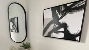 Une salle de bain avec un lavabo blanc, un miroir et un tableau abstrait noir accroché au mur.