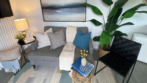 A modern living room with a grey sofa, blue and patterned cushions, a large green plant, a black chair, a framed artwork, and a small table with a lamp and a picture frame. - Unique container home  (El Cajon)