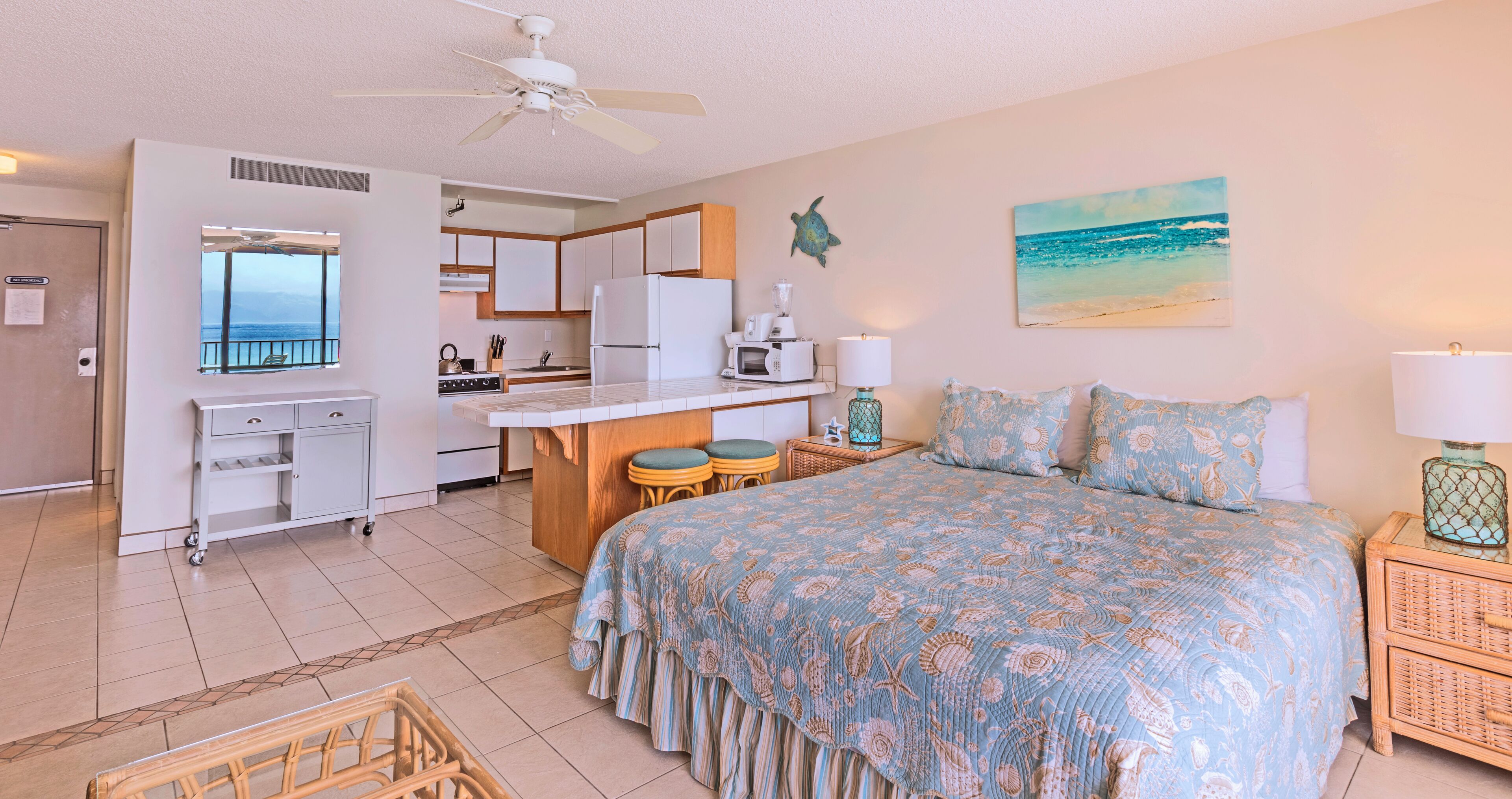A bedroom with a bed, a nightstand, a lamp, a window with a view of the ocean, a ceiling fan, and a kitchenette with a refrigerator, microwave, and sink.
