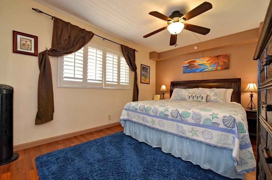 Un dormitorio con cama, ventilador de techo, ventana con cortinas, alfombra azul y un cuadro en la pared.