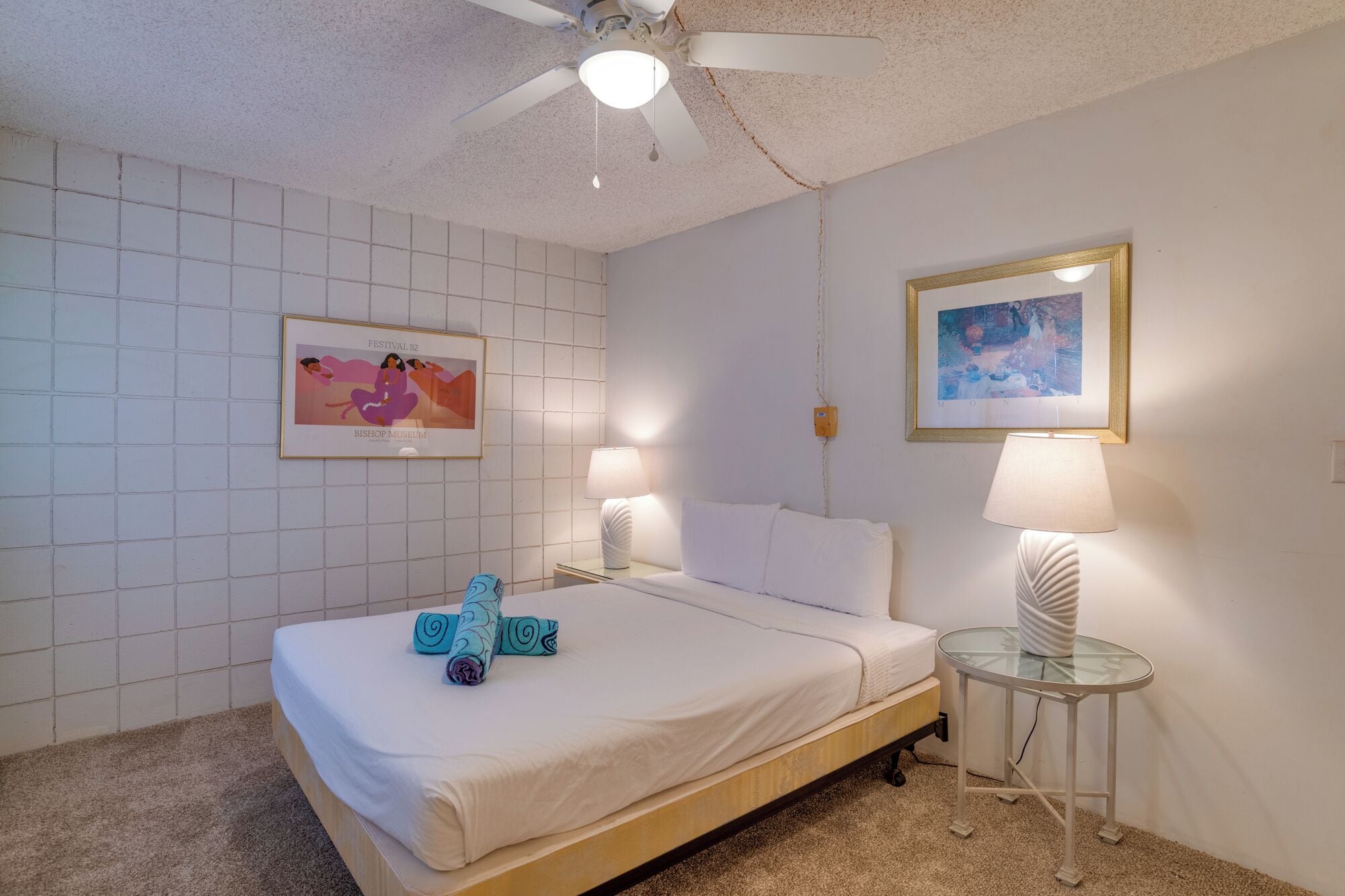 A neatly arranged hotel room with a bed, two bedside lamps, a ceiling fan, and two framed artworks on the walls.