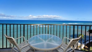Ein Balkon mit einem Glastisch und zwei Stühlen, der Blick auf einen Strand und das Meer freigibt.