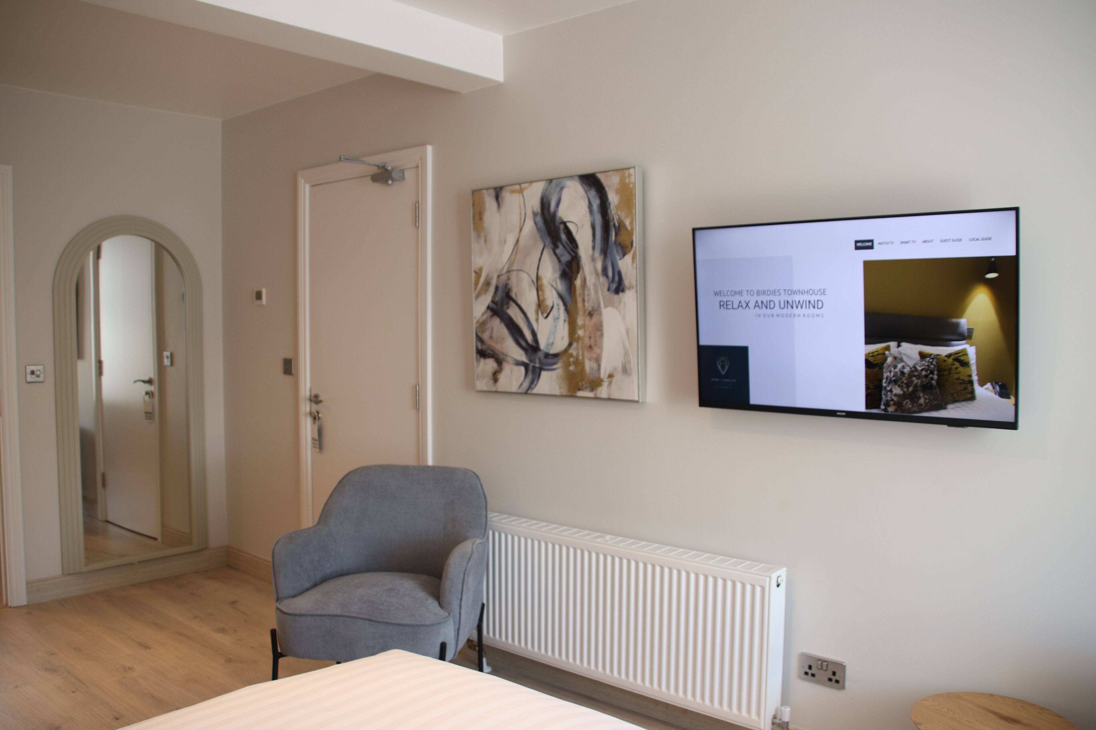 A modern hotel room with a grey armchair, a large abstract painting, and a television displaying a welcome message.