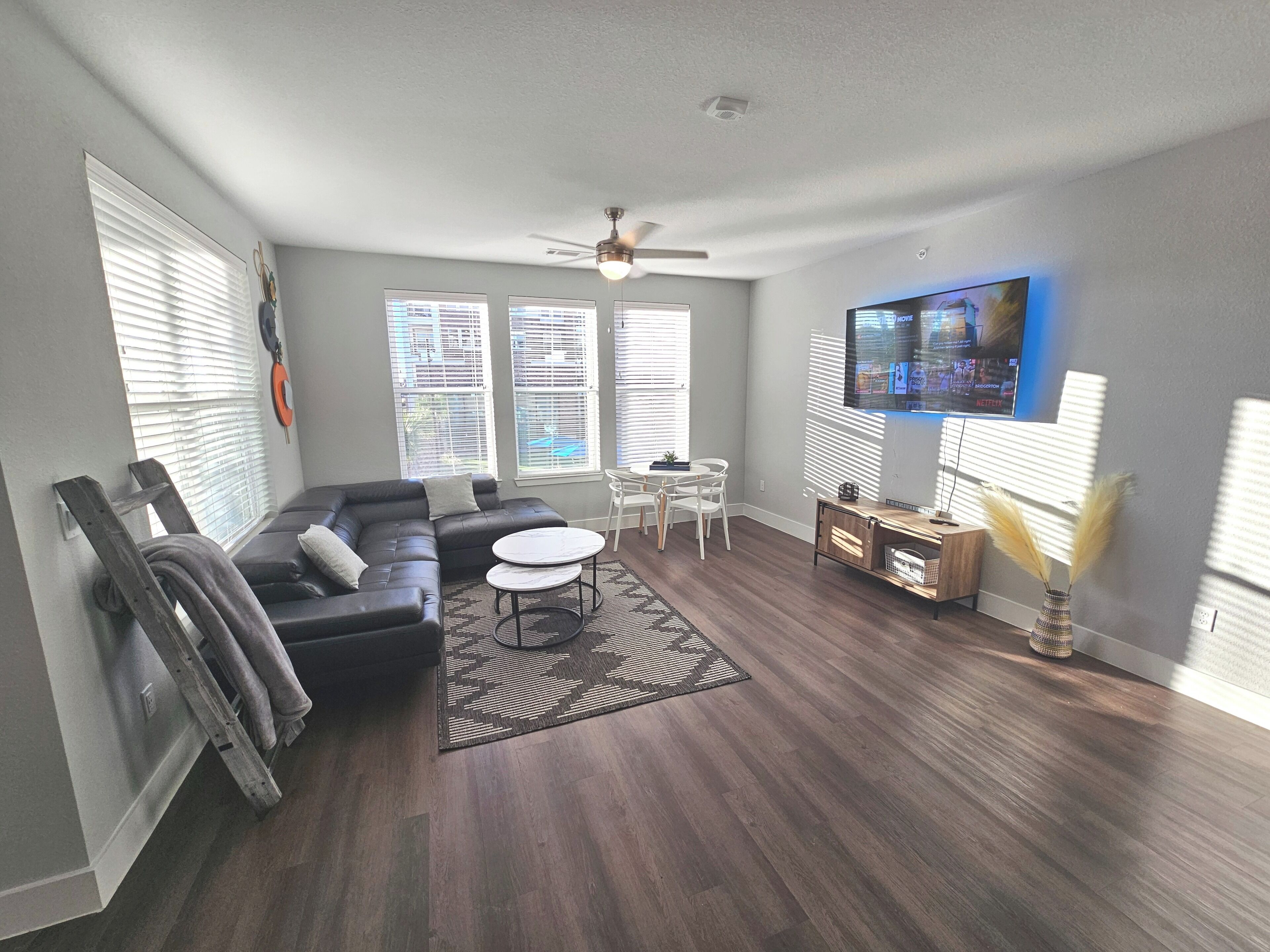 A modern living room with a sectional sofa, a dining table with chairs, a flat-screen TV mounted on the wall, and a ceiling fan.
