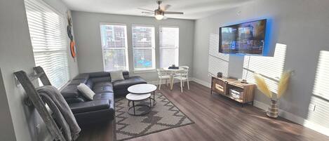 A modern living room with a sectional sofa, a dining table with chairs, a flat-screen TV mounted on the wall, and a ceiling fan.