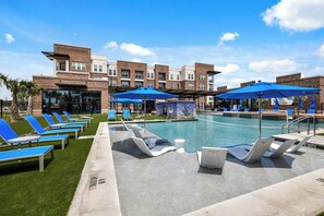 A pool area with lounge chairs and umbrellas, a brick building in the background, and a clear blue sky.
