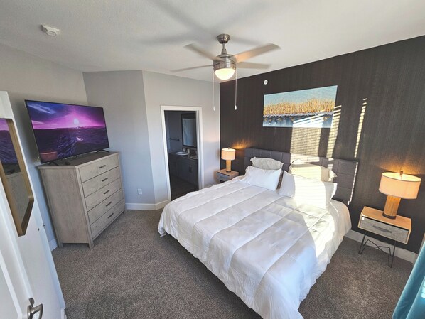 A bedroom with a bed, a TV, a dresser, a ceiling fan, and a painting on the wall.
