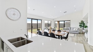 A modern kitchen with a large island, a dining area with a table and chairs, and a living room with a sofa and a plant.