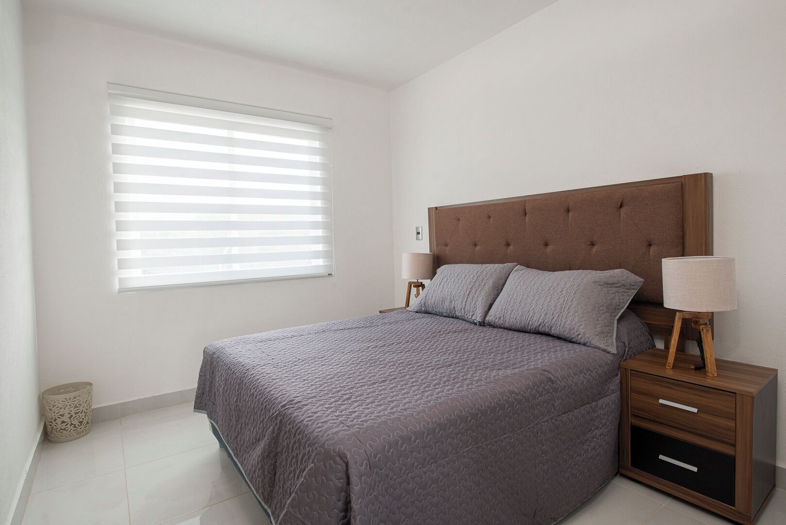 A bedroom with a large bed, a nightstand with a lamp, and a window with blinds.