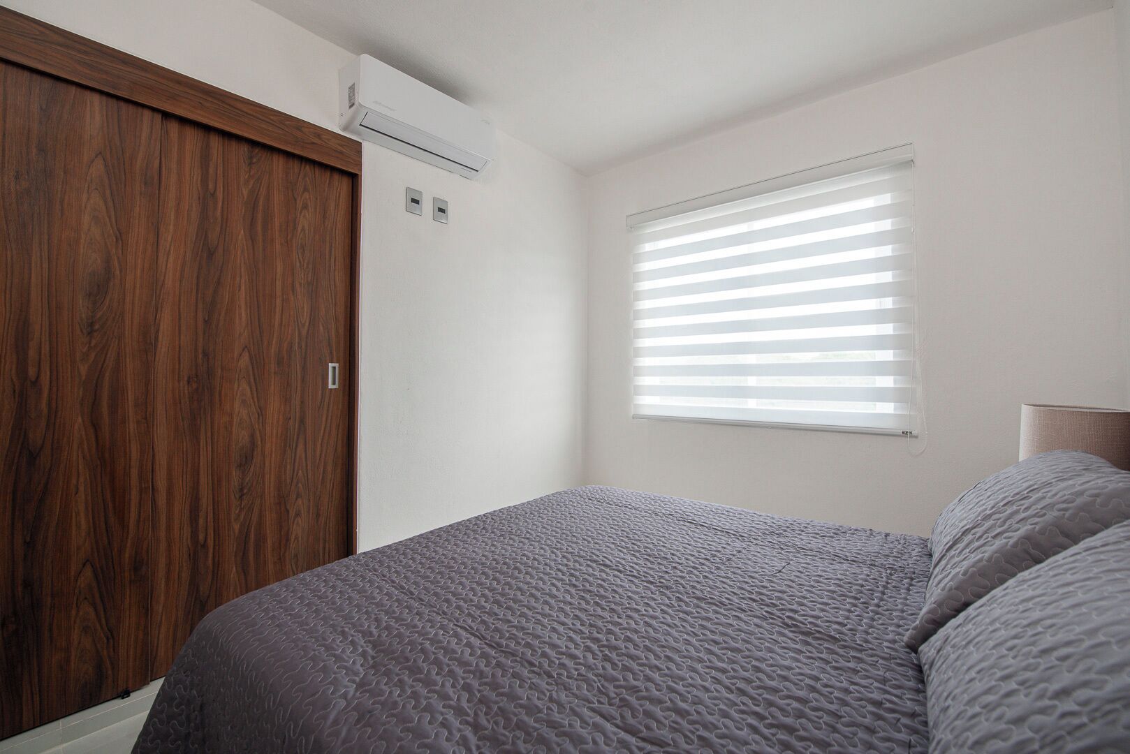 A bedroom with a bed, a wooden wardrobe, a window with blinds, and an air conditioning unit.