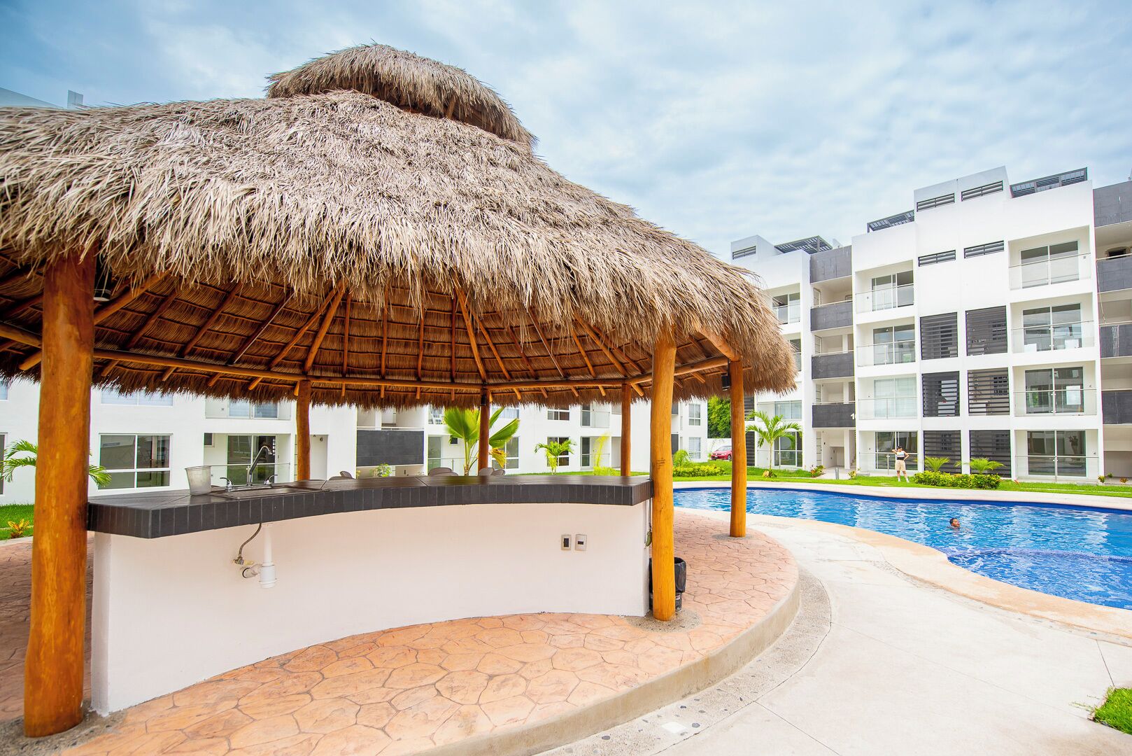 A poolside area with a thatched gazebo, a pool, and modern buildings in the background.
