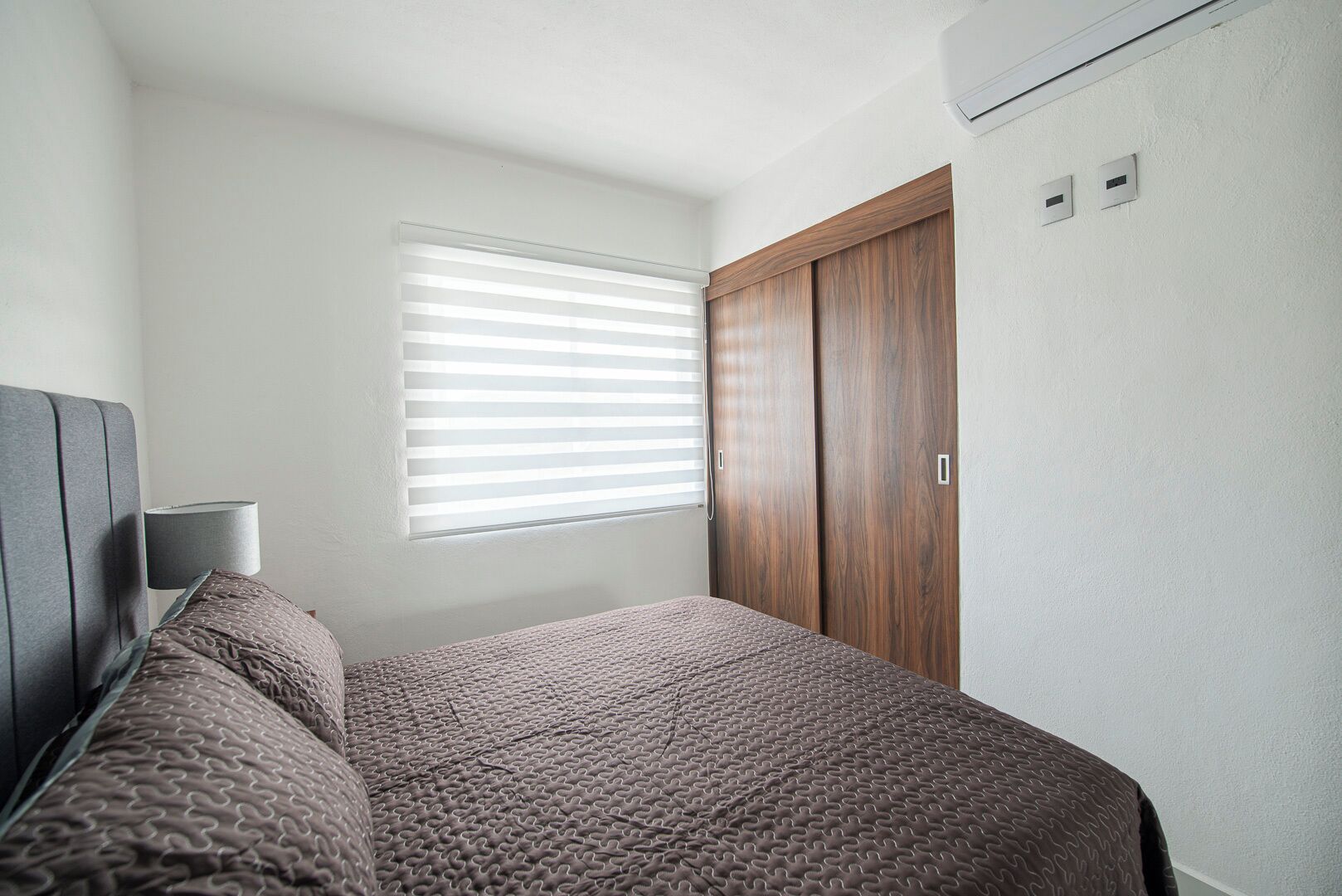 A bedroom with a bed, a window with blinds, a wardrobe, and a wall-mounted air conditioning unit.