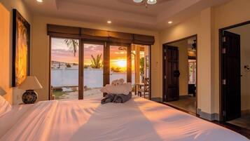 A hotel room with a bed, a nightstand, a lamp, a view of the sunset, and a door leading to another room.