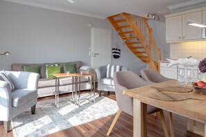 A modern living room with a staircase, grey sofas, a wooden dining table, and a kitchen area with white cabinets.