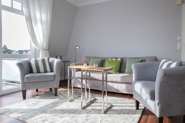 Ein modernes Wohnzimmer mit einem grauen Sofa, zwei Sesseln, einem Holztisch und einem großen Fenster mit weißen Vorhängen.