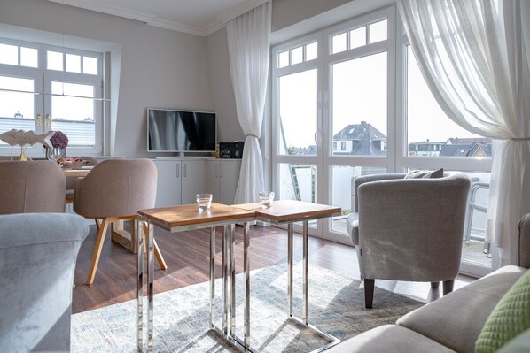 A modern living room with a wooden table, grey armchairs, and a sofa. A TV is mounted on the wall, and there is a view of buildings outside through large windows.