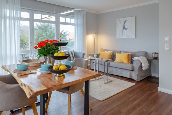 A modern living room with a dining area, featuring a wooden table, grey sofa, and a painting on the wall.