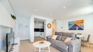 A modern living room with a grey sofa, a white coffee table, and a TV. There is a kitchen in the background with white cabinets and a black countertop.