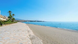 A cobblestone pathway leading to a sandy beach with clear blue water, palm trees, and buildings in the background.